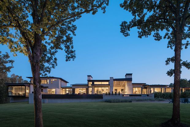 Wide exterior view of a large, low-slung contemporary residence at dusk, framed by mature trees and set behind an expansive manicured lawn. The home has a strongly horizontal composition, flat and shallow roof planes, extensive illuminated glazing, dark window frames, pale rendered walls, and a central recessed entry or courtyard-like zone.