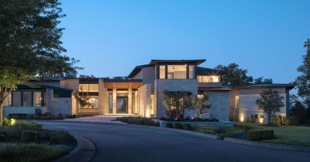 A dusk exterior view of a large contemporary single-storey residence with a layered, low-pitched roofline, pale masonry walls, extensive warm-lit glazing, landscaped garden beds, and an attached garage. The home is approached by a broad curved driveway and surrounded by mature trees and manicured planting.