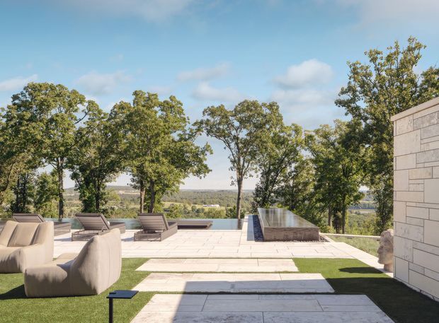 A spacious landscaped outdoor terrace overlooking a wooded valley, with pale stone paving, clipped lawn strips, sculptural lounge seating, sun loungers, and a long raised reflecting pool or water feature. A light stone-clad building wall frames the right side of the image. The scene has a clean, contemporary resort-like character with bright natural light and distant rural scenery.