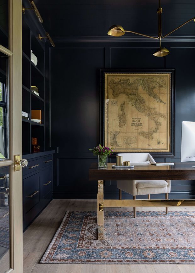 A dark, refined home office with deep navy paneled walls and matching built-in shelving and cabinetry. A substantial timber desk with brass-toned legs faces the room, paired with a light upholstered desk chair. A large framed antique-style map provides a focal point, while a sculptural brass multi-arm pendant adds warm contrast. The room features pale timber flooring and a large muted Persian-style rug, with daylight entering through the glazed door and window at left.