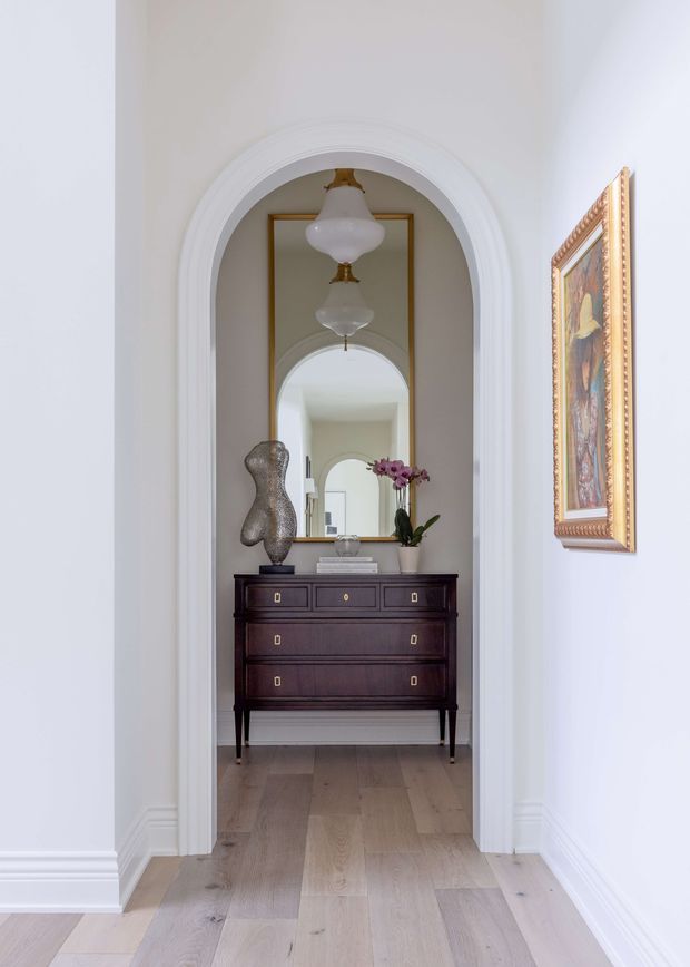 A bright, refined residential hallway or entry passage framed by a large white-painted arched opening with layered architrave detailing. A dark wood antique-style chest of drawers sits at the far end beneath a tall gold-framed mirror, creating a repeated arch reflection. The space has pale wide-plank timber flooring, white walls and decorative skirting, traditional pendant lights, a sculptural vase, orchid, books, and framed artwork. The composition feels elegant and symmetrical with classic detailing and a calm neutral palette.
