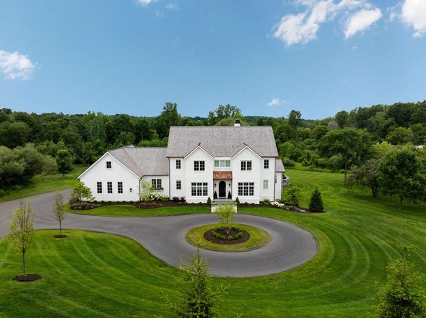 Aerial exterior view of a large white multi-gabled house set within expansive, carefully landscaped green grounds. The symmetrical main facade has multiple-pane windows, a central entrance, steep pitched gables, and grey shingle roofing. A circular driveway with a planted central island curves around the front of the property, with mature trees and open lawns surrounding the home under a clear blue sky.