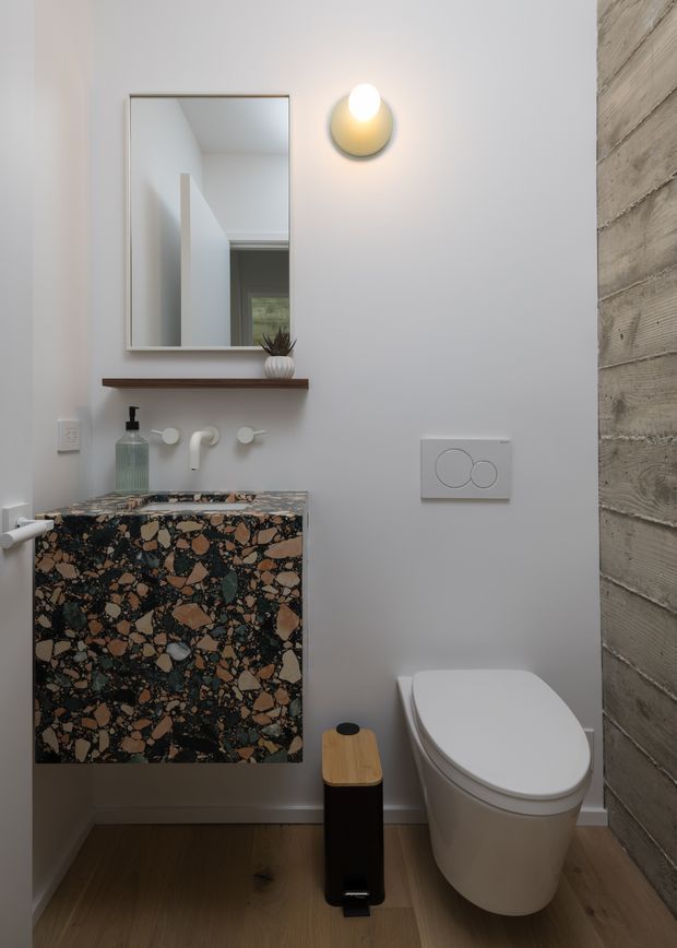 A compact contemporary bathroom with a wall-mounted terrazzo vanity, integrated basin and minimalist white wall-mounted tapware. A frameless rectangular mirror sits above a timber shelf, accompanied by a rounded wall sconce. The room includes a wall-hung white toilet, flush plate, timber-look flooring, and a textured horizontal timber feature wall.