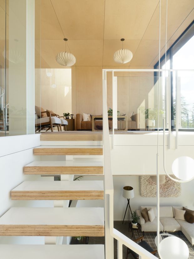 Bright contemporary interior featuring a floating timber staircase with white risers and clear glass balustrades. The stair connects a lower sitting area with an upper living room, which has warm plywood wall and ceiling panels, neutral upholstered seating, indoor plants, and sculptural white pendant lights. Floor-to-ceiling glazing brings in abundant daylight, while the open void creates a strong sense of vertical connection between levels.