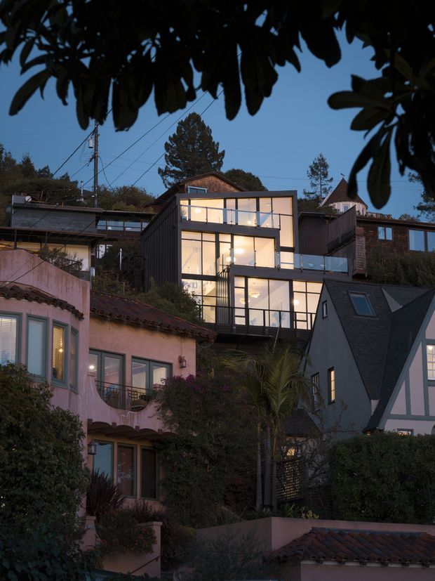 Dusk exterior view across a dense, leafy residential hillside, framed by dark tree foliage. The main subject is a narrow multi-storey contemporary home with a dark vertical-clad side wall and large black-framed floor-to-ceiling glazing, warmly illuminated from within. Surrounding homes include a pink stucco house with terracotta roof tiles and a steep-roofed Tudor-influenced house, creating a layered suburban neighbourhood setting.