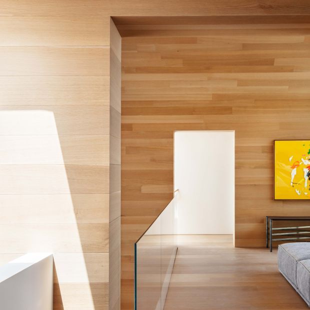 A refined contemporary interior corner featuring extensive warm timber wall and ceiling cladding, a white sculptural stair or partition element, and a clear glass balustrade. A partial grey upholstered sofa, metal-framed side table, and bright yellow artwork are visible at the right, while strong sunlight creates geometric highlights across the pale timber surfaces.