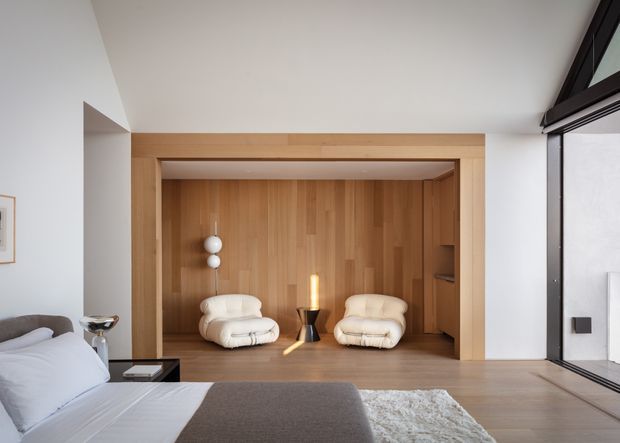 A serene contemporary bedroom with a low-profile bed in the foreground, opening onto a compact timber-lined sitting alcove. Two sculptural cream lounge chairs, a small dark side table, pendant light, and a freestanding candle-like light create a warm minimalist retreat. Full-height black-framed glazing is visible on the right, and the room uses pale timber flooring, white walls, and integrated timber joinery.