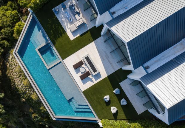 Aerial view of a contemporary residential property arranged around a long angular swimming pool. Several blue-grey clad buildings with pale metal pitched roofs surround pale paved terraces, outdoor lounge furniture, manicured lawn, and sculptural outdoor seating. Dense landscaping borders the site.