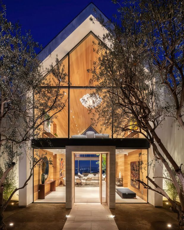 Night-time exterior view of a dramatic multi-storey residence framed by mature trees. The facade combines large black-framed glazing with warm vertical timber wall cladding, a steep triangular gable form, and a projecting dark horizontal balcony or canopy. Through the central entry, a refined interior and distant illuminated view are visible. The landscaped approach includes paved flooring and integrated ground lighting.