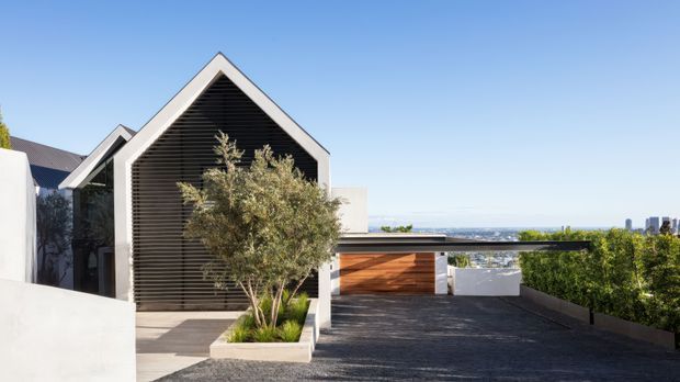 Exterior view of a contemporary residential property with a prominent steep gabled entry volume, dark horizontal cladding, white rendered walls, and a flat-roofed garage or carport extending to the right. A paved driveway and raised planter lead toward the entrance, with a distant urban skyline visible beyond the property.
