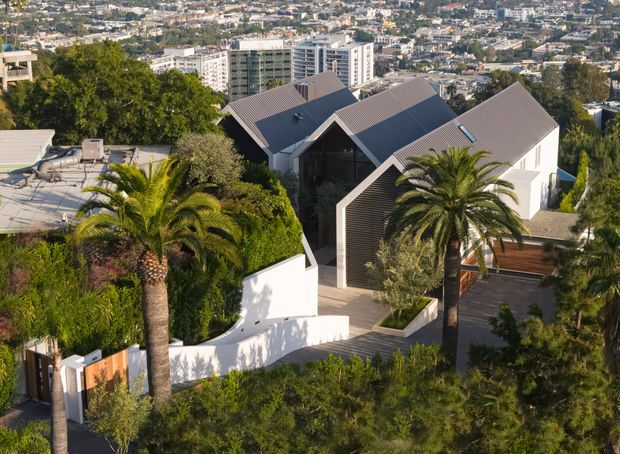 Elevated exterior view of a contemporary multi-volume residence set on a steep, landscaped urban site. The home features several pitched metal roofs, white rendered walls, dark vertical cladding, extensive glazing, terraces, and mature palm planting, with a dense cityscape visible beyond.