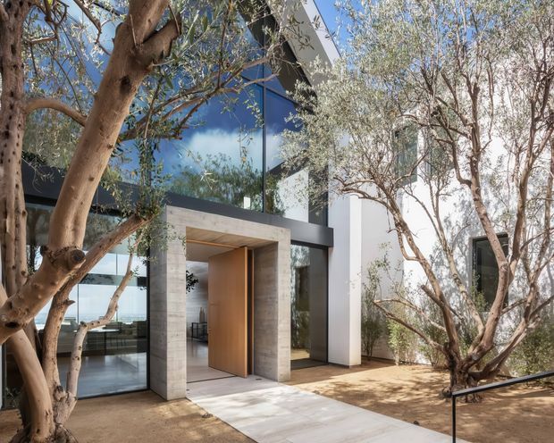 Exterior view of a contemporary two-storey home entrance, framed by mature olive-like trees and a landscaped courtyard. The facade combines large reflective glazing, dark structural framing, white rendered walls, and a substantial timber pivot door set within a pale stone or concrete portal. A paved walkway leads to the recessed entry.
