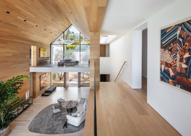 Contemporary multi-level residential interior viewed from an upper landing, with extensive light timber wall and ceiling lining, matching timber flooring, a double-height living area, large glazed openings, sculptural seating, and a stair hall. A colourful framed artwork provides an accent against the white walls.