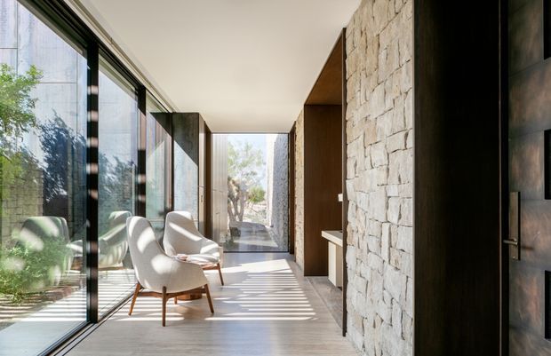 A refined contemporary entry hall or transitional corridor with full-height black-framed glazing on the left, two sculptural lounge chairs, pale timber or stone-look flooring, and a striking rough-cut light stone feature wall on the right. Dark timber built-in joinery and a substantial dark door frame the passage, while strong daylight creates linear shadows across the floor.