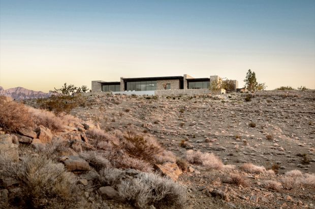 A low, elongated modern residence set on a barren rocky desert hillside. The building has strong horizontal lines, dark flat roof planes, extensive floor-to-ceiling glazing, and a restrained stone-toned exterior that blends with the rugged arid landscape. Sparse desert shrubs and distant mountains surround the property under a clear sky.