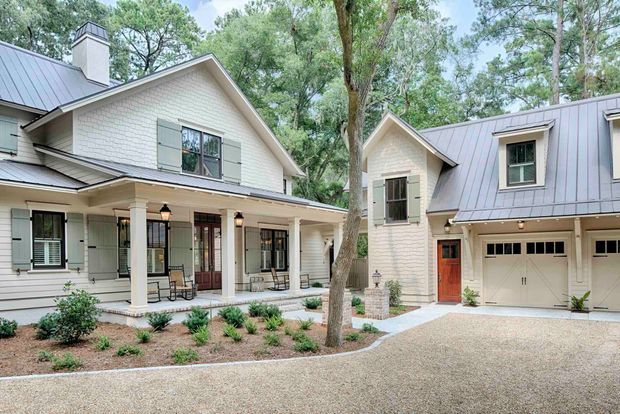 Exterior view of a light-coloured, multi-wing residence set among mature trees. The home features white painted brick walls, grey standing-seam metal gable roofs, shuttered windows, covered front verandah with square columns, and an attached garage with a separate entry door. Gravel or exposed-aggregate driveway and restrained front landscaping complete the scene.
