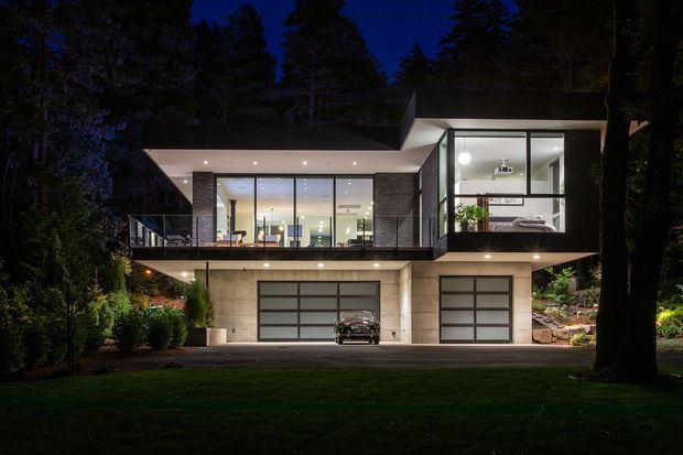 Night-time exterior photograph of a contemporary two-storey house set among mature trees. The home has a broad cantilevered upper level, extensive floor-to-ceiling glazing, dark brick or masonry wall sections, pale concrete or rendered lower walls, a balcony terrace, and two integrated garage doors. Warm interior lighting reveals open living spaces and a bedroom through the glazing.