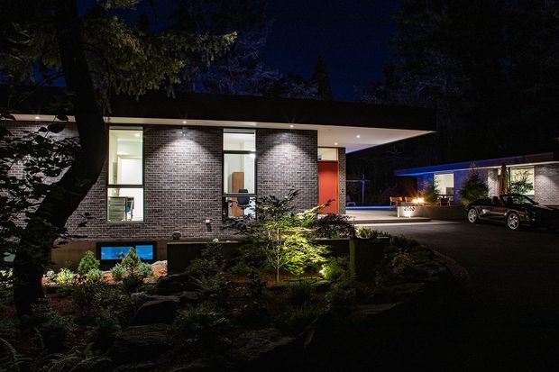 Night-time exterior view of a low, single-storey modern residence with a broad flat roof, dark brick feature walls, extensive vertical glazing, and a vivid red entry door. Landscaped planting and illuminated garden beds frame the foreground, while a driveway and partially visible detached structure sit to the right.