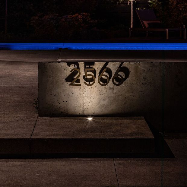 A close-up nighttime view of a minimalist concrete address marker bearing the number 2566, set into a paved entry area. Small ground lighting illuminates the textured concrete, with a blue-lit swimming pool and dark garden seating visible in the background.