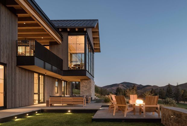 Contemporary multi-level residence at dusk, featuring dark vertical timber cladding, extensive floor-to-ceiling glazing, a projecting upper balcony with black metal balustrade, exposed timber soffits, and a low-pitched tiled roof. A stone-clad wall and timber deck create an outdoor seating area with armchairs and a fire feature, overlooking a landscaped setting and distant hills.