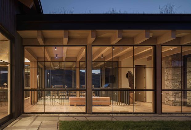 Twilight exterior view of a contemporary pavilion-like building with extensive black-framed floor-to-ceiling glazing. Warm timber soffits and exposed ceiling beams shelter a covered terrace, while a substantial natural stone wall forms part of the right side. Interior seating, timber joinery, and a dining or living area are visible through the glass, with a paved path and lawn in the foreground.