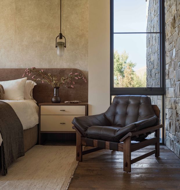 A warm, refined bedroom corner featuring a partially visible bed with a rounded upholstered headboard, layered neutral bedding, a simple two-drawer bedside table, and a vintage-style dark leather lounge chair. The room combines textured plaster walls, a full-height dark-framed window, a rugged natural stone feature wall, and dark timber flooring. A suspended pendant light and understated floral branch add subtle character.