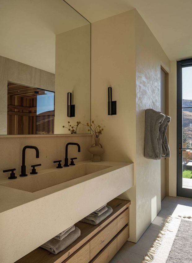 Warm contemporary bathroom with a long double vanity, integrated pale stone or solid-surface basins, matte black wall-mounted and basin tapware, a large frameless mirror, and light timber drawer cabinetry. Textured beige plaster walls, simple black vertical sconces, folded towels, and a full-height glazed door create a refined, natural interior with views outside.