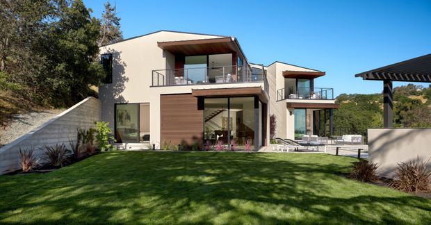 Exterior view of a contemporary multi-level residential home set into a landscaped sloping site. The facade combines smooth light rendered walls, large glazed openings, warm timber garage or entry doors, projecting flat roof eaves, and metal balcony balustrades. A paved outdoor area and separate covered structure are visible to the right, with lawn and established trees surrounding the property.