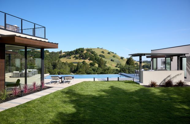 A modern residential outdoor setting with a manicured lawn, swimming pool, and hillside views. The left side shows a glass-walled living area with a broad timber-edged roof terrace and black metal balustrade, while the right side contains a separate flat-roofed pavilion with extensive glazing. A paved path connects the buildings and pool area, with sparse ornamental planting around the edges.