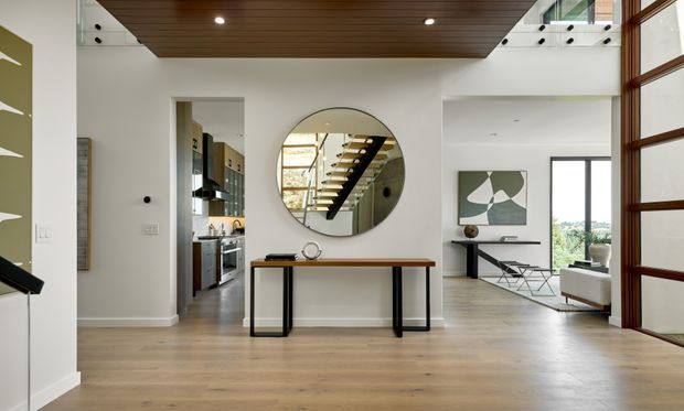 Bright contemporary open-plan entry and living area with pale timber flooring, white walls, a dark timber-lined ceiling feature, and a slim console table beneath a large circular mirror. The mirror reflects a floating stairway with timber treads and a glass balustrade. An open kitchen is visible to the left, while the living room with artwork and floor-to-ceiling glazing is visible to the right. Tall timber-framed glazing and a double-height void contribute to the spacious, refined interior.