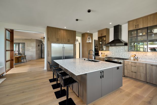 A spacious contemporary kitchen with a large marble-look island, integrated sink and seating for several people. Warm timber cabinetry extends across the rear wall and around a stainless-steel refrigerator, with open shelving and glass-front storage. A black rangehood, dark fixtures and pendant lights provide contrast against the pale stone splashback and light timber flooring. The kitchen opens to an adjoining dining or living area through timber-framed glazing and wide circulation space.