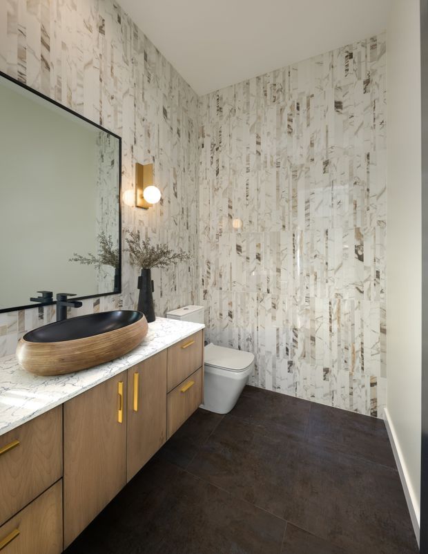 A contemporary bathroom featuring a floating timber vanity with gold-toned handles, a white veined stone benchtop, black vessel basin and tapware, large black-framed mirror, and wall sconces. The walls are clad in a vertically patterned light marble-look tile, complemented by dark floor tiles and a white toilet.