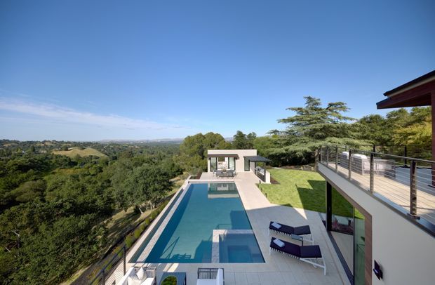 Elevated exterior view of a contemporary multi-level residence with a long rectangular swimming pool, paved pool terrace, outdoor lounge furniture, glass-and-metal balustrades, and expansive landscaped views over a leafy suburban or semi-rural setting. The home has clean horizontal lines, extensive glazing, and a flat-roofed pool pavilion.
