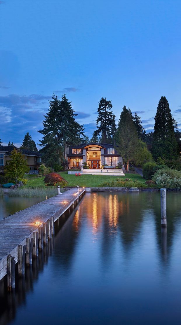 A dusk exterior view of a large waterfront house, photographed across a calm body of water. The home has expansive illuminated glazing, a prominent central entrance, a curved upper roof feature, and broad steps leading to a landscaped lawn. A timber jetty extends from the foreground toward the property, with tall evergreen trees surrounding the site and warm interior lighting reflected in the water.