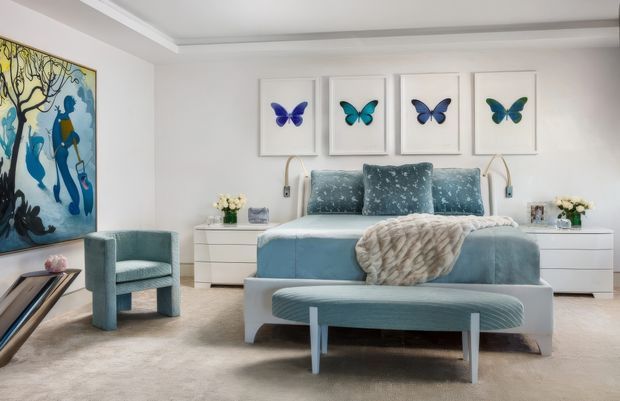 Bright, refined bedroom with a centered upholstered bed in pale blue, matching blue bedside tables and a curved bench at the foot. Four framed butterfly artworks hang above the bed, while a large colourful artwork, teal accent chair, bedside lamps, flowers and a soft neutral carpet complete the room. The ceiling features layered recessed trim and the overall styling is elegant and contemporary with playful natural motifs.
