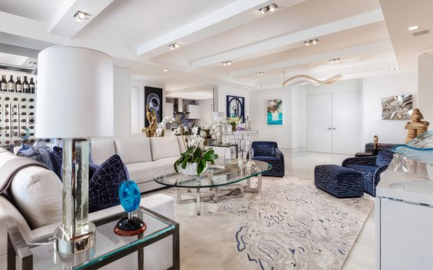Bright, spacious open-plan living room with a predominantly white interior, sculptural recessed ceiling details and track lighting. The room features a large cream sectional sofa, navy upholstered lounge chairs and ottomans, a transparent glass coffee table with a chrome or metal base, mirrored and glass side tables, white built-in cabinetry, decorative artwork, and a large abstract-patterned area rug. A bar or bottle display is visible at the rear, and the overall styling is polished and glamorous.