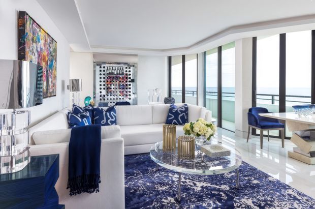 Bright, spacious contemporary living room with expansive floor-to-ceiling glazing overlooking a waterfront or ocean horizon. The room features white sofas, bold navy patterned cushions and rug, a glass coffee table, sculptural metallic accessories, blue accent furniture, and colourful wall art. A dining area is visible at the right, while a wine display or bar feature appears in the background.