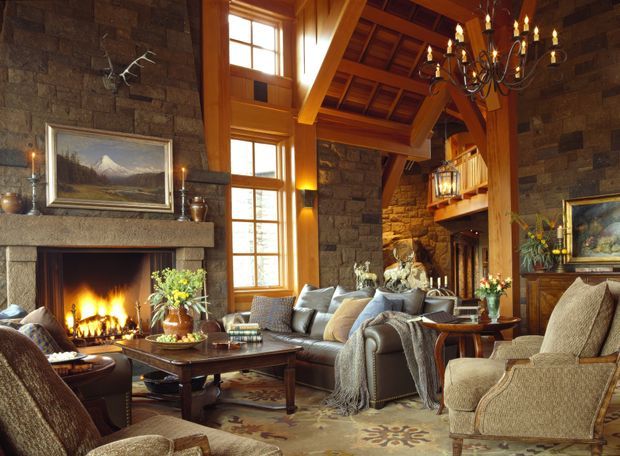 A warm, rustic living room with a large stone fireplace, exposed timber beams and vaulted timber ceiling, tall divided-light windows, a mezzanine balcony, and a traditional chandelier. Upholstered seating, a substantial timber coffee table, framed landscape art, and layered textiles create a cozy lodge-like atmosphere.