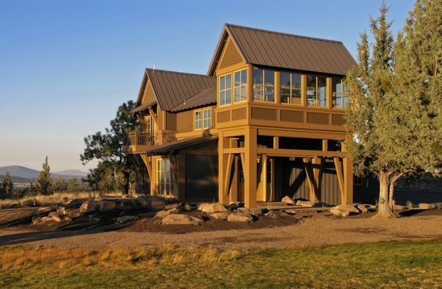 Exterior view of a tall timber-framed house in a dry, open landscape. The building has multiple pitched gable roofs with dark metal roofing, warm ochre-brown timber cladding and exposed structural posts, and extensive upper-level glazing. A covered lower level and balcony-like projecting volumes create a raised, multi-storey form.