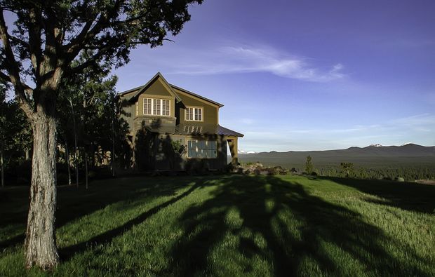 A two-storey detached house set in a spacious grassy rural landscape, viewed from outdoors. The home has a prominent pitched gable roof, muted olive-grey exterior walls, white-trimmed multi-pane windows, and a small covered entry or porch. Mature trees frame the foreground, while distant forested hills and mountains extend across the background.