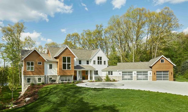 A large detached suburban residence set within a landscaped, wooded site. The house has multiple pitched gabled roof volumes, a light exterior with warm timber-clad gable accents, tall multi-pane windows, and a covered front entry. A separate matching garage with two garage doors sits to the right, connected by a paved driveway and circular forecourt.