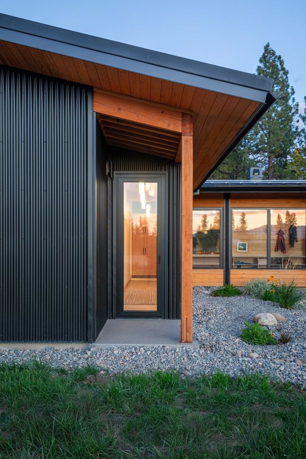 Exterior view of a modern single-storey building entrance and courtyard. The facade combines dark vertically ribbed cladding with warm timber soffits, exposed timber posts and extensive glazing. A narrow glazed door sits beneath a deep timber-lined roof overhang, opening toward a gravelled landscaped courtyard with low planting. The composition highlights the roof edge, sheltered entry and material contrast.