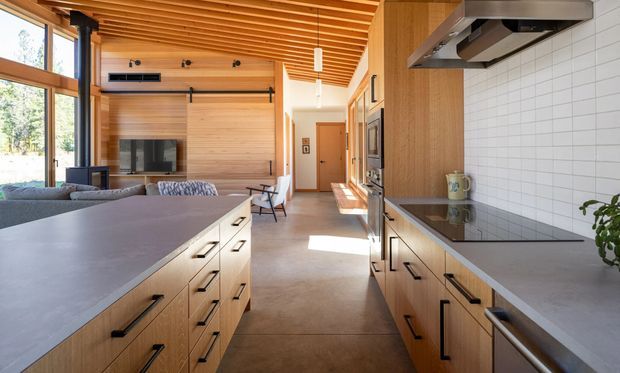 A bright open-plan kitchen viewed from the cooking area, with a large central island, extensive light timber cabinetry, dark linear handles, grey benchtops, integrated appliances and a white tiled splashback. The kitchen opens directly to a timber-lined living room with a fireplace and large windows. A slatted timber ceiling and warm natural materials create a contemporary, refined interior.