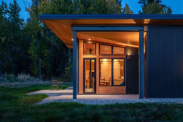 A compact modern pavilion or small cabin set within a wooded landscape, photographed at dusk. The building has a strong flat projecting roof with warm timber soffits, dark metal structural framing, extensive timber-framed glazing, and a vertically ribbed dark exterior wall. Warm interior lighting reveals a small furnished room, while a paved path and outdoor seating lead toward the entrance.