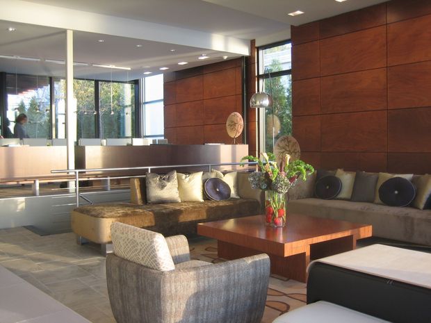 A spacious, refined contemporary living room or lobby with a large sectional sofa, upholstered lounge chair, and substantial square timber coffee table. The room features extensive dark timber wall panelling, full-height glazing, mirrored surfaces, a pale tiled floor, recessed ceiling lighting, and metallic pendant lights. Neutral upholstery and cushions are accented by a floral arrangement on the coffee table.