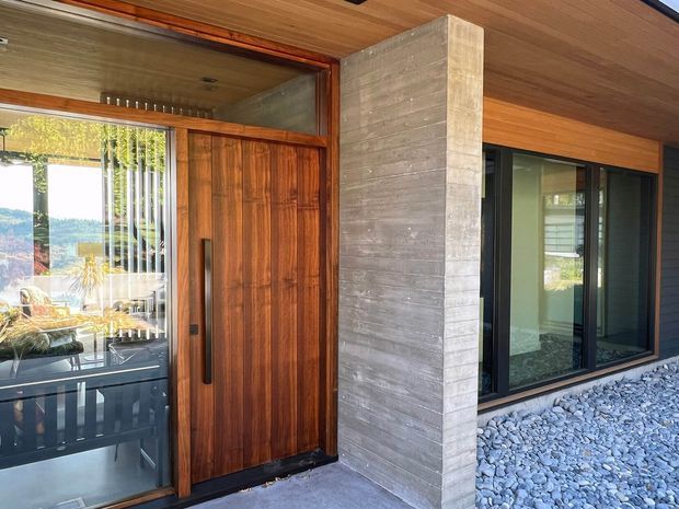 Close-up exterior view of a contemporary home entry, featuring a tall natural timber pivot door, exposed board-formed concrete column, dark-framed glazing, timber soffit, and a gravel-covered ground surface. Interior furnishings and a scenic landscape are visible through the glazing and reflections.