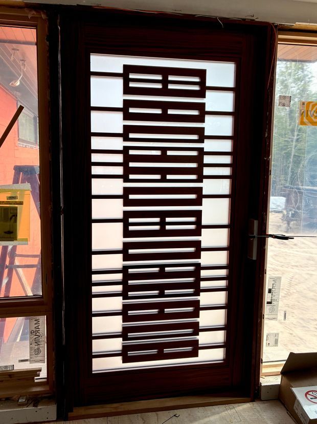 Close-up view of a contemporary glazed entry door with a dark timber-toned frame and decorative geometric metal grille. Frosted or translucent glass panels provide privacy while admitting light. Adjacent glazed sidelights and unfinished construction surroundings are visible, suggesting the door is being viewed in a building or showroom under construction.