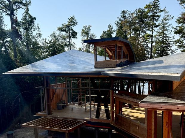 Exterior view of a contemporary multi-level timber-framed building under construction, set among mature trees. The structure has broad low-pitched roof planes, exposed timber framing, extensive open glazing areas, and a raised sheltered upper-level balcony or pavilion. Construction materials and unfinished openings are visible throughout.