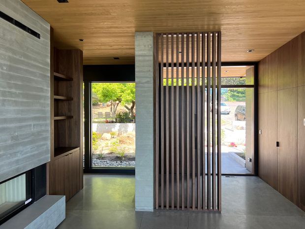 A refined contemporary interior entry space with a polished concrete floor, warm timber-lined ceiling and wall panelling, a raw concrete feature wall and bench, and a dark timber vertical slatted screen dividing the circulation area. Built-in timber shelving and a low horizontal window are visible on the left, while full-height glazing and an external doorway bring in natural light on the right.