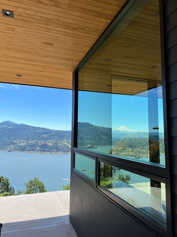 A modern glazed corner beside a covered outdoor terrace, featuring expansive black-framed windows, dark exterior cladding, and a warm timber-lined soffit or ceiling. The glazing reflects and reveals a lake, wooded hills, and a distant snow-capped mountain, creating a strong indoor-outdoor connection and panoramic alpine waterfront outlook.